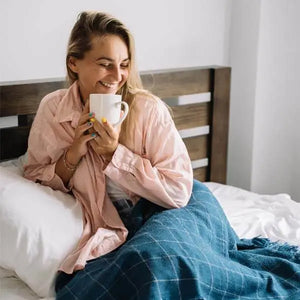 Une femme souriante tenant une tasse dans ses mains allongée sous un plaid à carreaux bleu